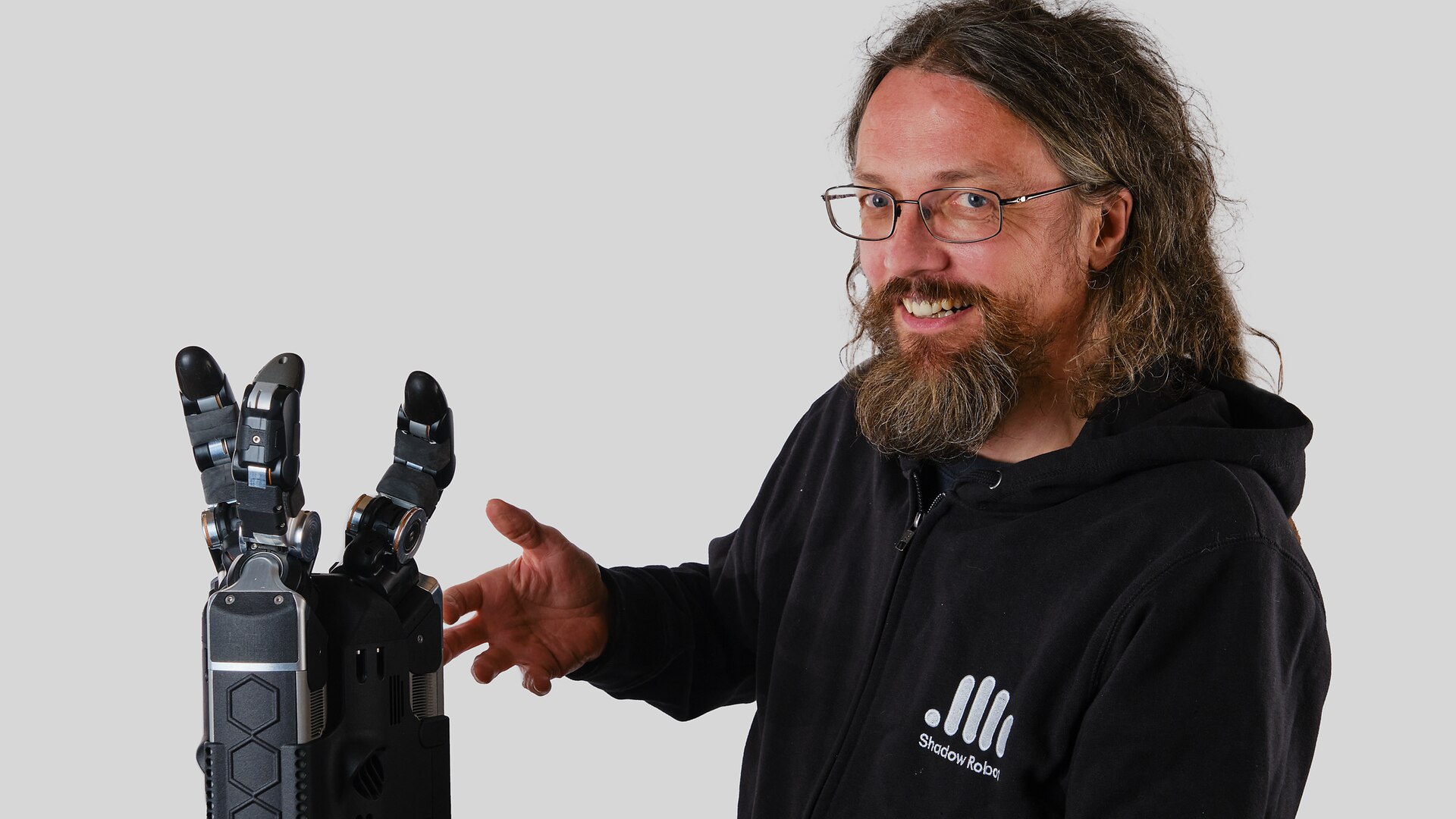 A man with long hair and a beard holds a robotic arm, showcasing a blend of human and technological innovation.